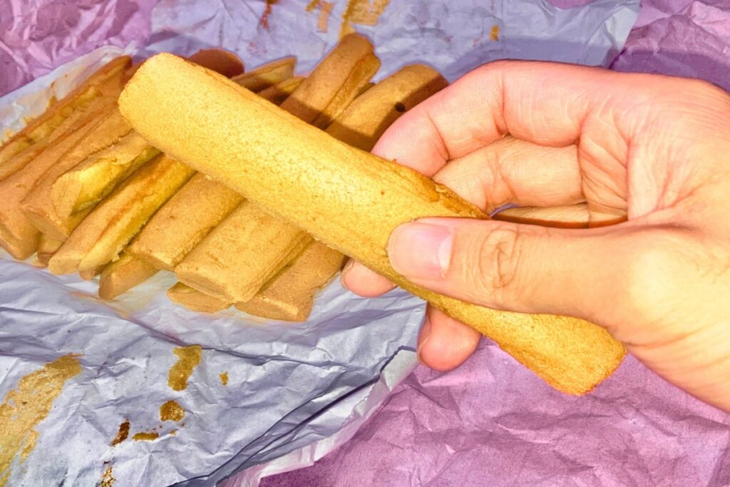 ピンクと白の包み紙に載った茶色いお菓子をバックに、右手で1本だけお菓子を持っています。