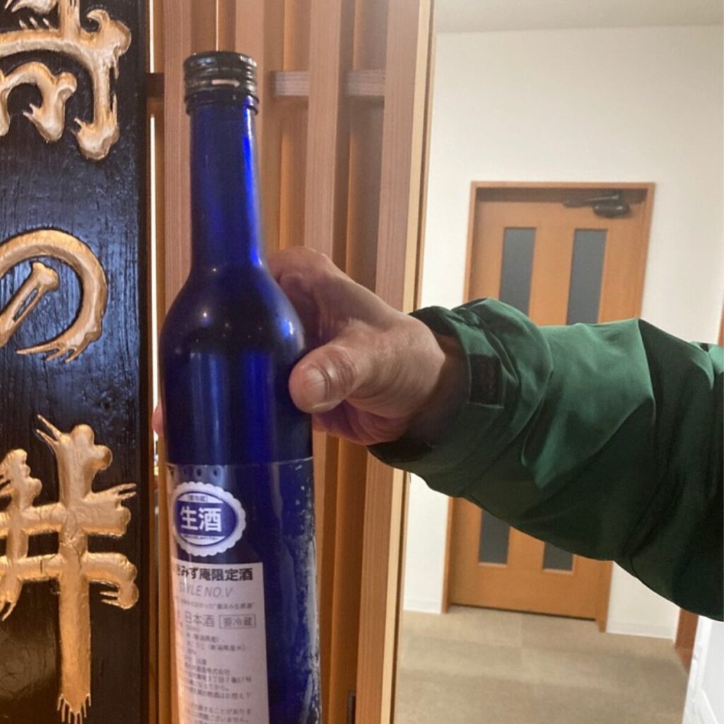 「生酒」と書かれたラベルが貼ってある青い酒瓶を、緑色の服を着た人が持ち上げています。