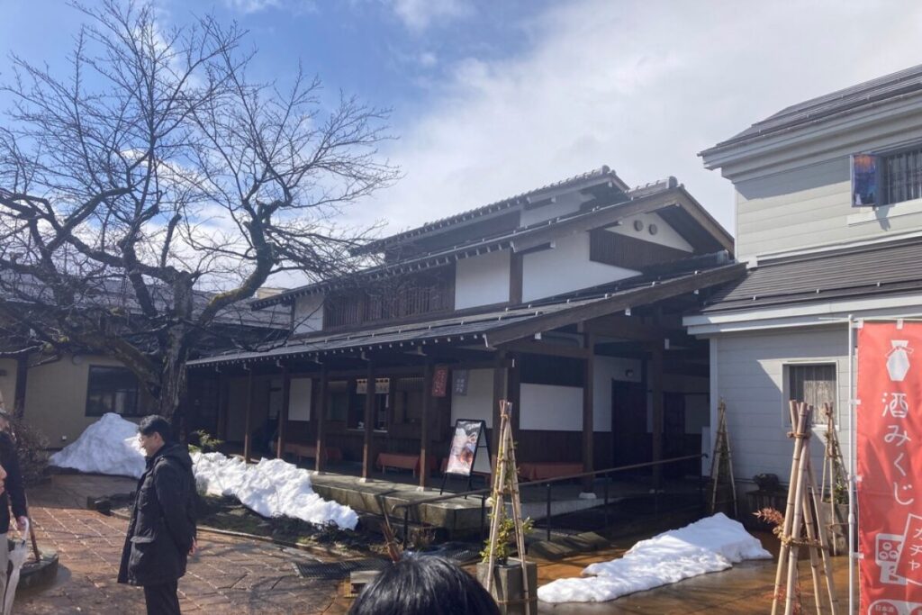 古民家が写っています。手前には木の生えた庭のような空間があり、雪が残っています。