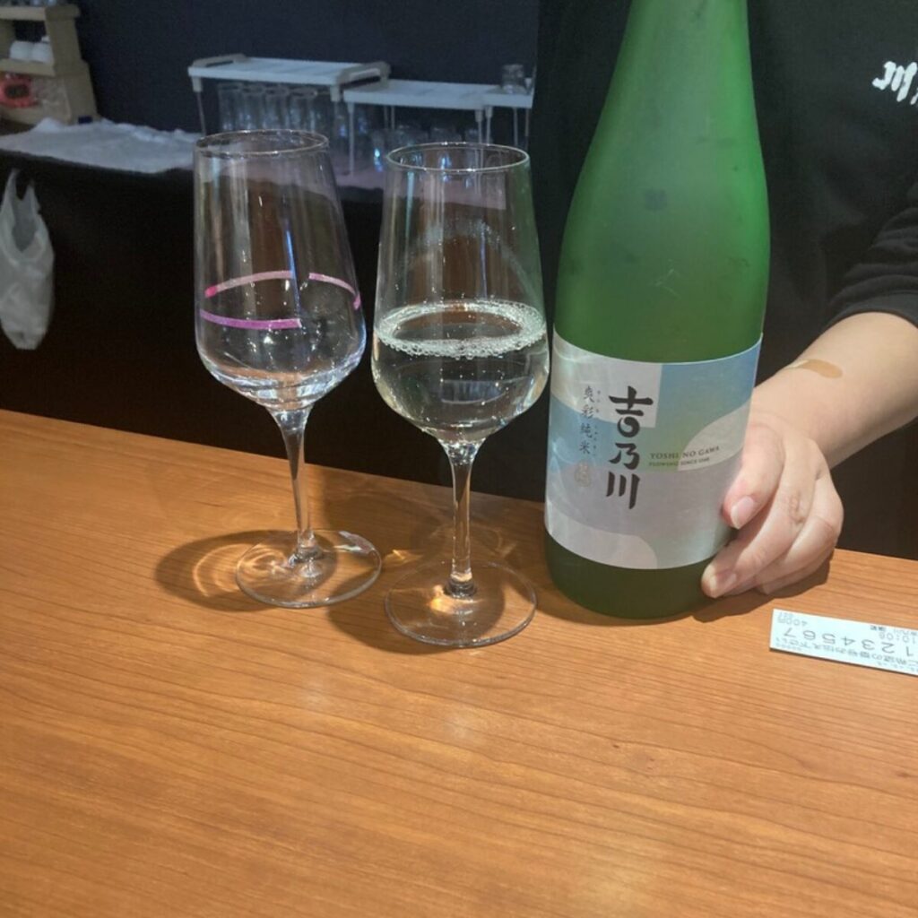 日本酒の入ったグラスが2つ並んでいて、酒瓶と人の手が写っています。
