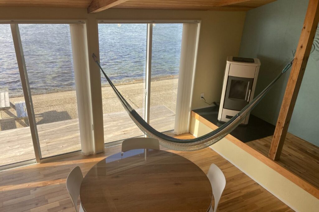 円形のガラステーブル越しに見る室内。窓際にはハンモックが設置され、右奥には薪ストーブ（またはペレットストーブ）があります。