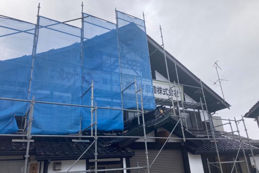 「高の井酒造株式会社」の看板がある建物の屋根に、鉄骨が組まれて安全ネットがかかっています。