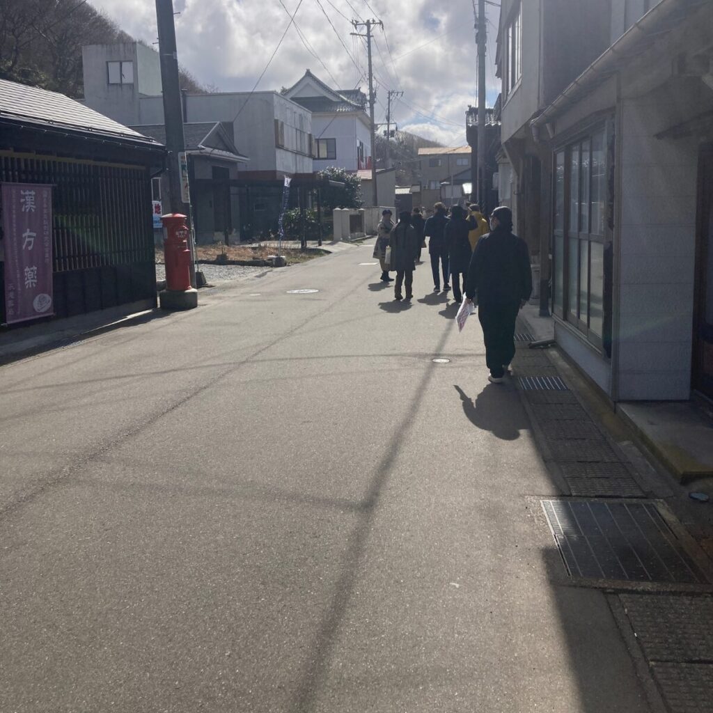 街中の風景を写した写真です。両側に家があり、中央よりやや右側に列になって歩く人たちが映っています。