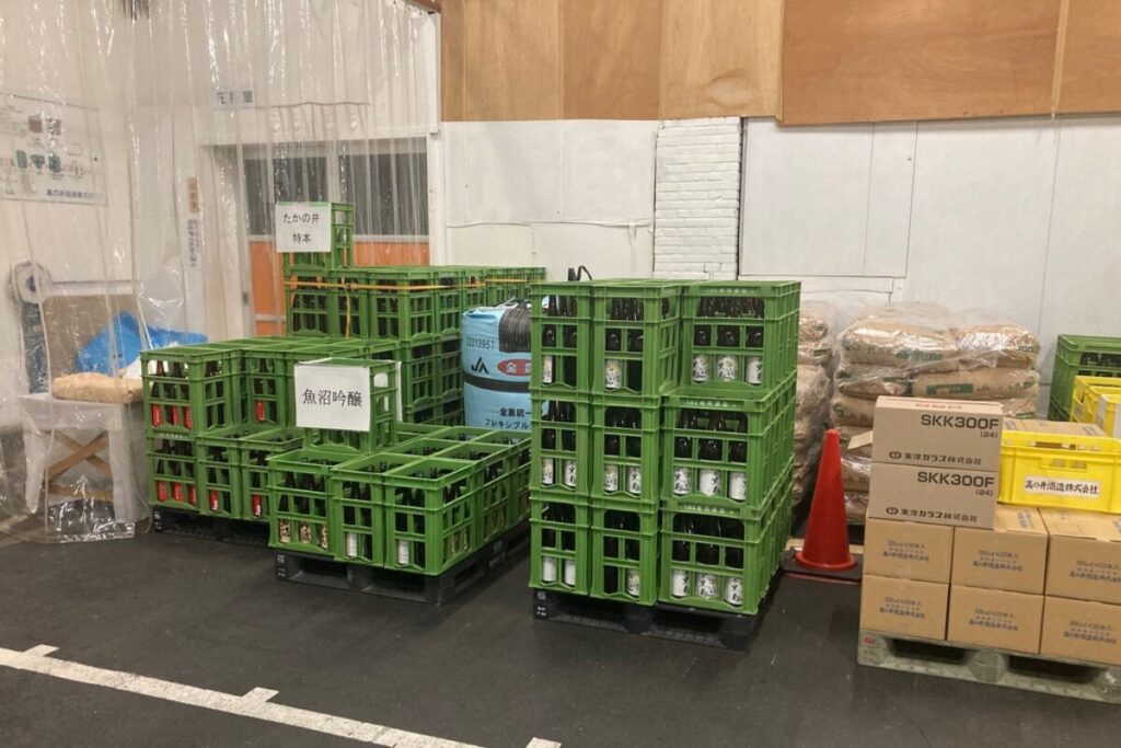 酒瓶がまとまっている緑色のケース、段ボールなどが置かれています。緑色のケースには「たかの井特本」「魚沼吟醸」などのラベルが貼られています。