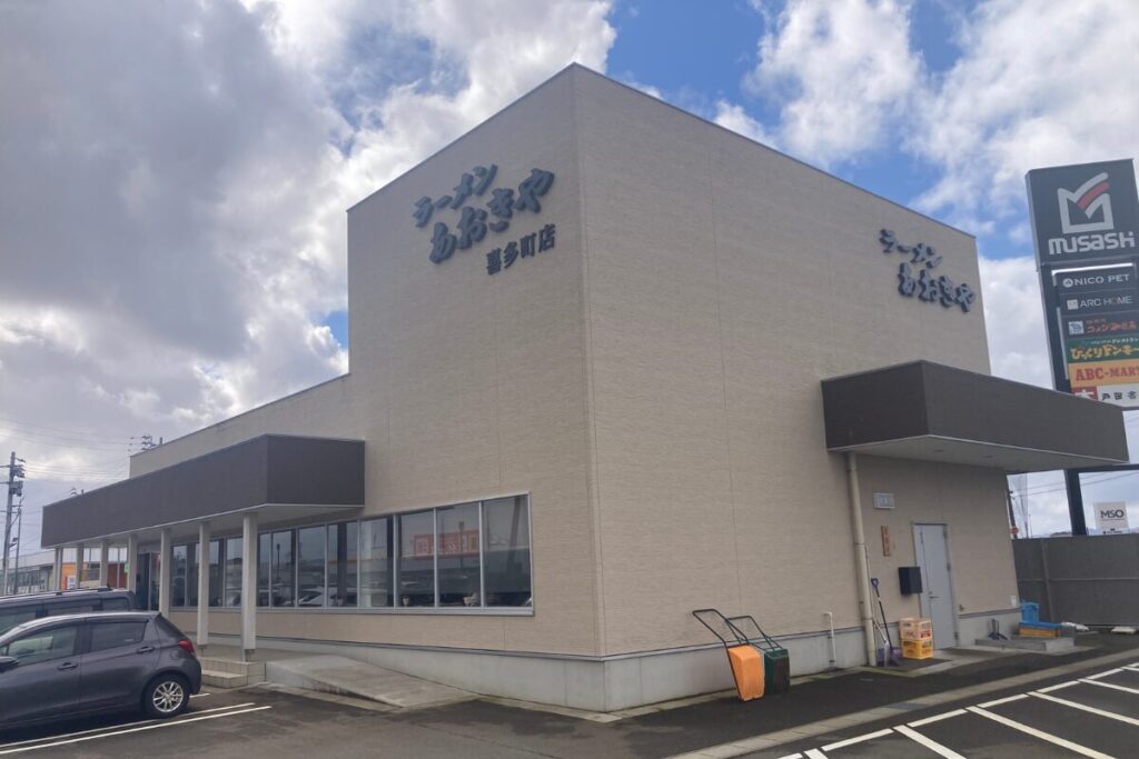 青空を背景に、ラーメン屋の店舗が大きく映っています。手前は駐車場で左側には車が見切れていて右側の奥には看板があります。