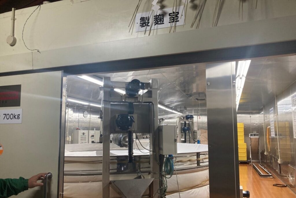 「製麹室」と書かれたプレートが部屋の入り口の上に貼ってあります。室内には円形の大きな機械があり、壁はステンレスです。