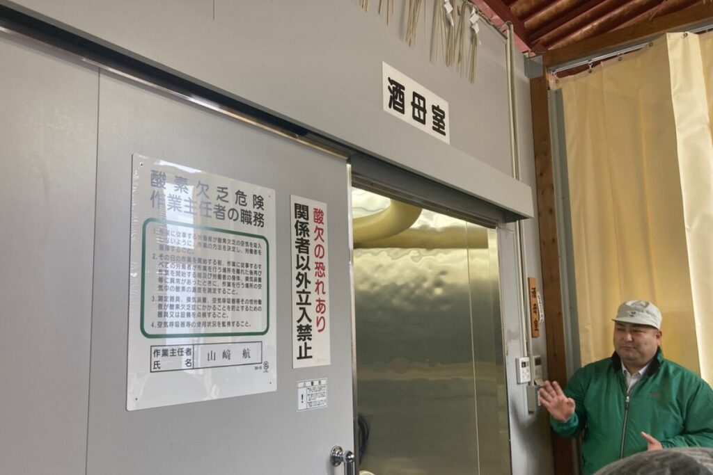 「酒母室」と書かれた部屋の入り口の前で、作業着の男性が手を広げて話をしています。