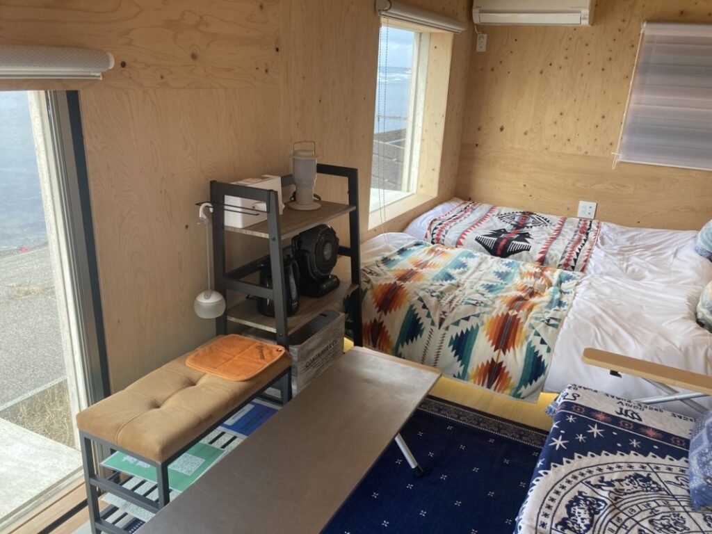 右側にシングルサイズのベッドが2つ、真ん中に3つの棚があり上段にランプとティッシュ箱、中段にサーキュレーター、下段に荷物入れがあります。