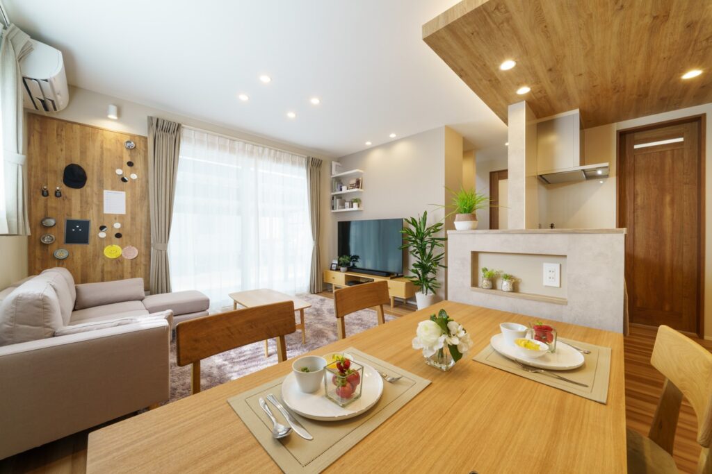 リビングが写っています。壁と天井は白く、ところどころ木製になっています。画面手前に木製のテーブルがあり、フルーツの入った皿が2つ、対角に置いてあります。画面中央、部屋の奥には膝ほどの高さにテレビがあり、それを視聴できる角度で低めのテーブルとソファが設置されています。