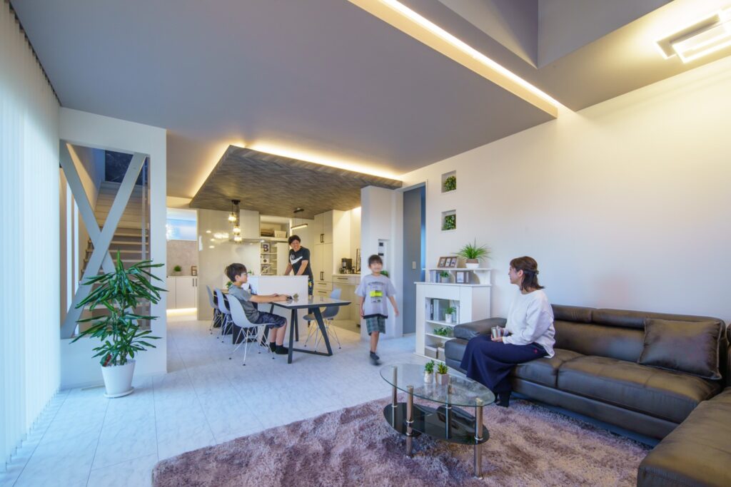 リビングで家族が団欒しています。画面右側の黒いソファに母親が座っており、画面中央には母親に向けて笑顔を浮かべて立っている子どもが1人、椅子に座っている子どもが1人います。その奥には父親が写っています。