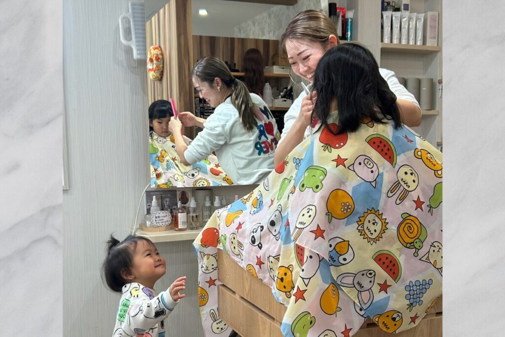 カット施術しながら笑顔の髙野さんと、施術の様子を笑顔で見ている子ども。