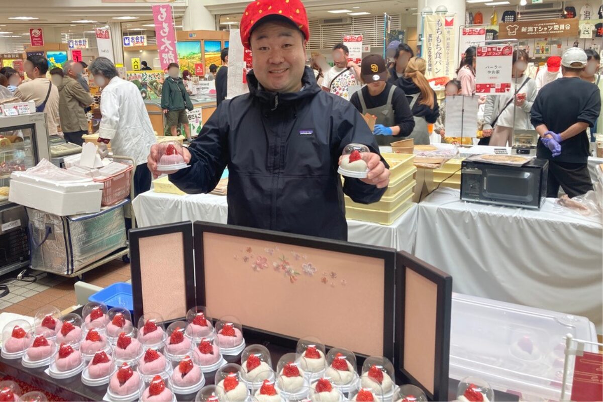 いちごを模した帽子を被った男性が、右手に赤色、左手に白色のいちご大福を持っています。